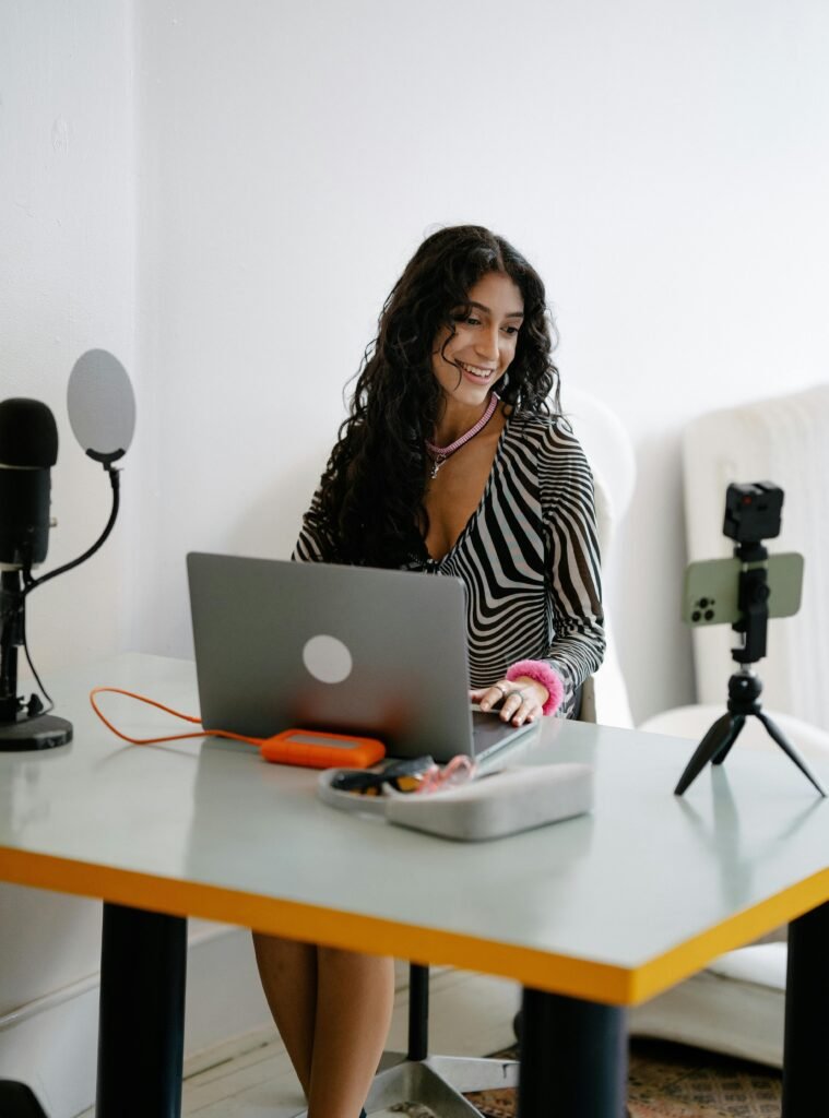 Smiling woman sits at desk using laptop, with camera and microphone setup for remote work.